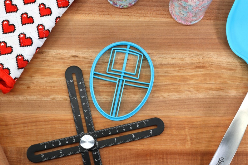May include: A blue plastic cookie cutter shaped like a circle with a square and lines inside. The cutter is on a wooden surface with a black ruler and a white and red fabric with hearts.