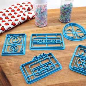 May include: Five blue plastic cookie cutters shaped like retro cassette tapes and a record player. The cutters are on a wooden surface with sprinkles in jars and a rolling pin.