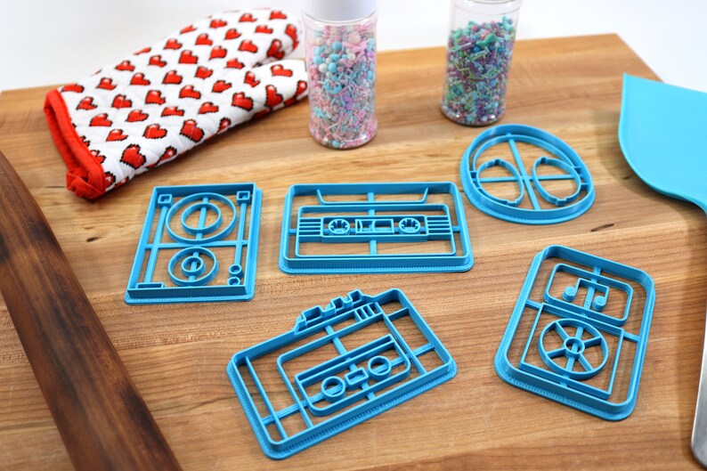 May include: Five blue plastic cookie cutters shaped like retro cassette tapes and a record player. The cutters are on a wooden surface with sprinkles in jars and a rolling pin.