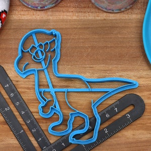 May include: Blue plastic dinosaur cookie cutter. The cutter is shaped like a dinosaur standing on two legs with its head tilted to the side. The cutter is on a wooden surface.