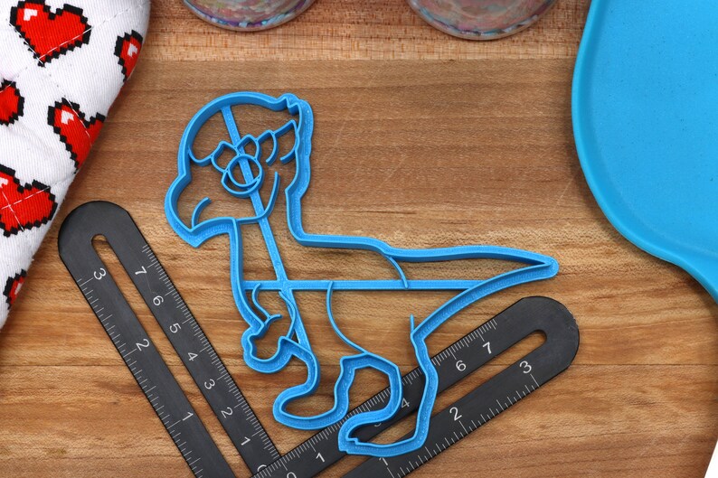 May include: Blue plastic dinosaur cookie cutter. The cutter is shaped like a dinosaur standing on two legs with its head tilted to the side. The cutter is on a wooden surface.