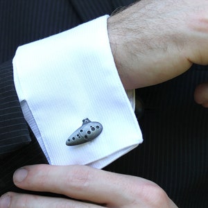 May include: A pair of cufflinks shaped like a silver ocarina, a musical instrument.