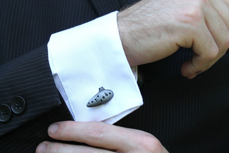 May include: A pair of cufflinks shaped like a silver ocarina, a musical instrument.