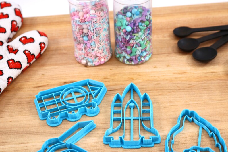 May include: Blue plastic cookie cutters shaped like a house, rocket, and a dinosaur on a wooden surface. Two jars of colorful sprinkles are in the background.