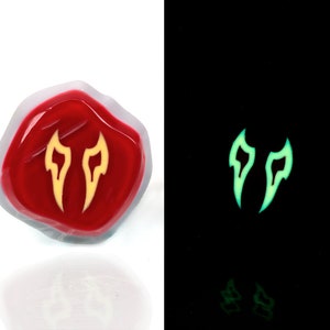May include: A red resin dice with a yellow glow-in-the-dark symbol of two flames. The dice is shown in both normal and dark lighting conditions.