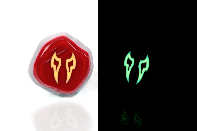 May include: A red resin dice with a yellow glow-in-the-dark symbol of two flames. The dice is shown in both normal and dark lighting conditions.
