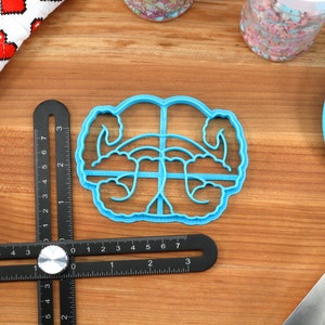 May include: A blue plastic cookie cutter shaped like a pair of hands holding a heart. The cookie cutter is on a wooden surface with a ruler and a spatula.