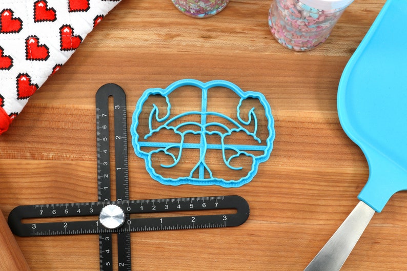 May include: A blue plastic cookie cutter shaped like a pair of hands holding a heart. The cookie cutter is on a wooden surface with a ruler and a spatula.
