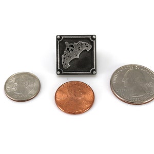 May include: A square, silver-toned metal pendant with an ornate design, flanked by a dime, a penny, and a quarter. The pendant features a detailed, stylized floral or leaf motif within a square frame. The coins are arranged around the pendant.