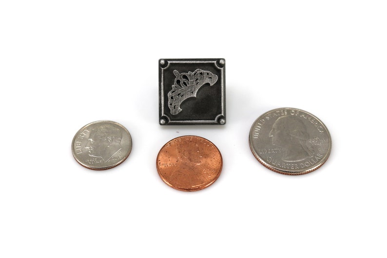 May include: A square, silver-toned metal pendant with an ornate design, flanked by a dime, a penny, and a quarter. The pendant features a detailed, stylized floral or leaf motif within a square frame. The coins are arranged around the pendant.