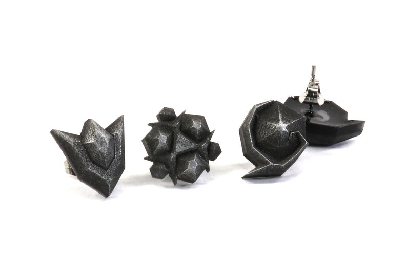 May include: Three black geometric earrings with a textured finish. The earrings are in the shape of a point, a cluster of points, and a curved shape.