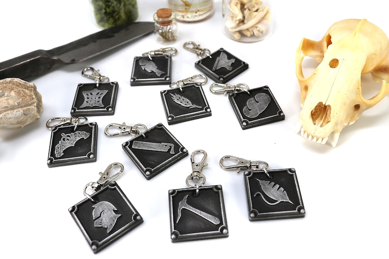 May include: Ten silver keychains with different symbols on each. The symbols include a fish, a feather, a cloud, a crown, a leaf, a hammer, a helmet, a knife, a crescent moon, and a sword. The keychains are on a white surface.