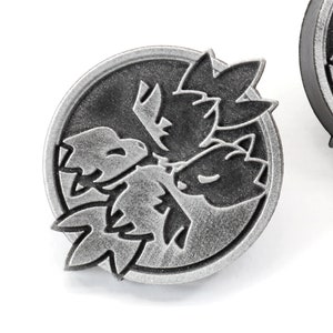 Op de afbeelding: Een zilverkleurige metalen pin met een zwart-wit ontwerp van drie gestileerde bladeren. De pin is rond en heeft een getextureerd oppervlak.
