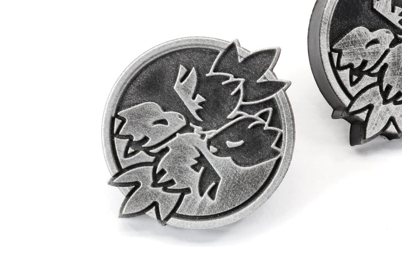 Op de afbeelding: Een zilverkleurige metalen pin met een zwart-wit ontwerp van drie gestileerde bladeren. De pin is rond en heeft een getextureerd oppervlak.
