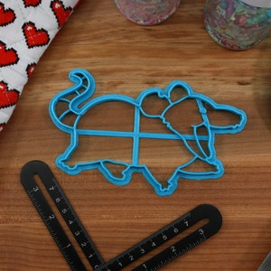May include: A blue plastic cookie cutter shaped like a rat. The cutter is on a wooden surface with a ruler and a blue silicone spatula.