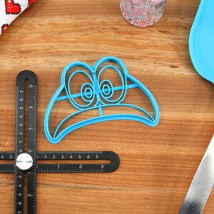 May include: Blue plastic cookie cutter shaped like a cartoon face with large eyes. The cutter is on a wooden surface.