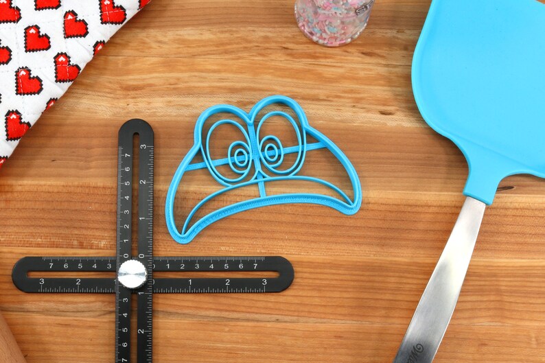 May include: Blue plastic cookie cutter shaped like a cartoon face with large eyes. The cutter is on a wooden surface.