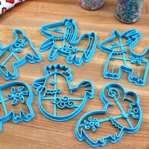 Puede incluir: Conjunto de siete cortadores de galletas de pl&aacute;stico azul con forma de varios animales, incluyendo un gallo, un cerdo, una cabra, un caballo y una oveja. Los cortadores est&aacute;n sobre una superficie de madera.