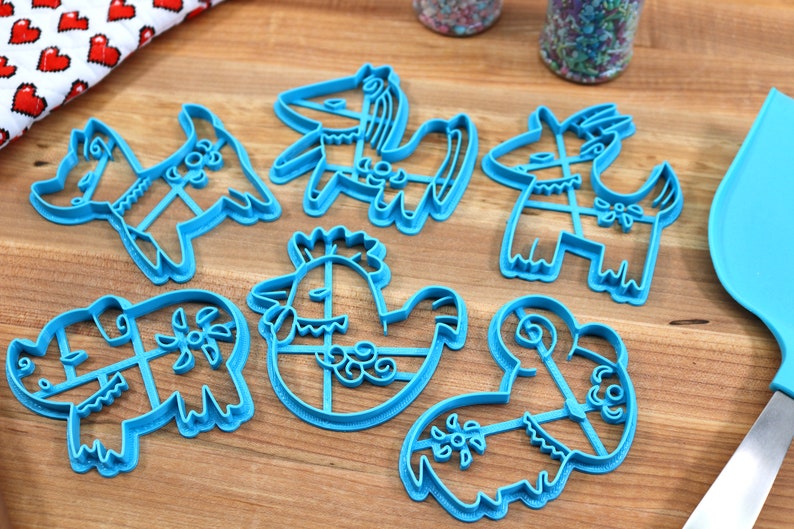 Puede incluir: Conjunto de siete cortadores de galletas de pl&aacute;stico azul con forma de varios animales, incluyendo un gallo, un cerdo, una cabra, un caballo y una oveja. Los cortadores est&aacute;n sobre una superficie de madera.