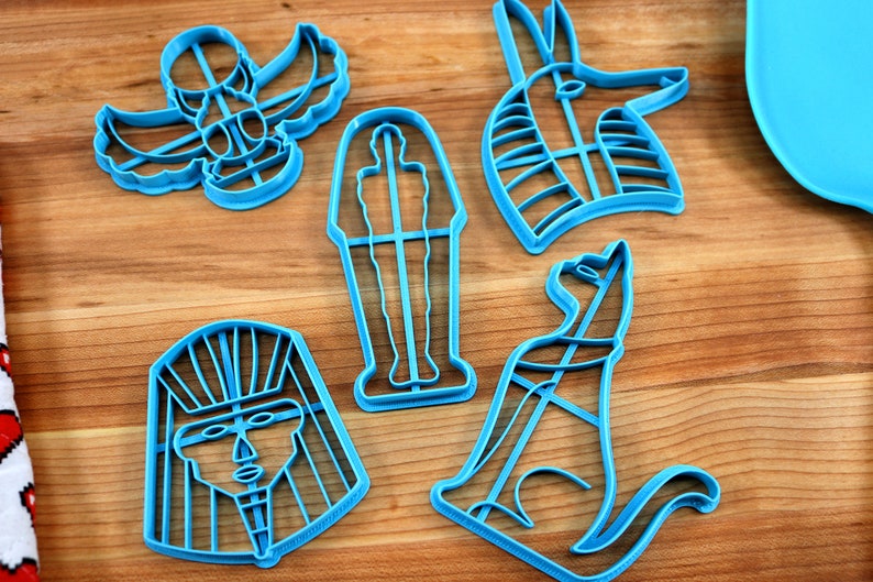 May include: A set of five blue plastic cookie cutters shaped like Egyptian symbols. The cutters include a scarab beetle, a pharaoh's head, a sarcophagus, a cat, and an ankh.