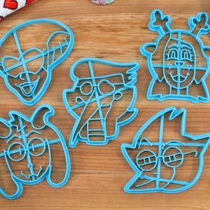Peut inclure: Cinq emporte-pi&egrave;ces en plastique bleu clair repr&eacute;sentant diff&eacute;rents personnages de dessins anim&eacute;s, dont un renne, un chien et un personnage avec des lunettes. Les emporte-pi&egrave;ces sont sur une surface en bois avec une spatule bleue et un rouleau &agrave; p&acirc;tisserie.