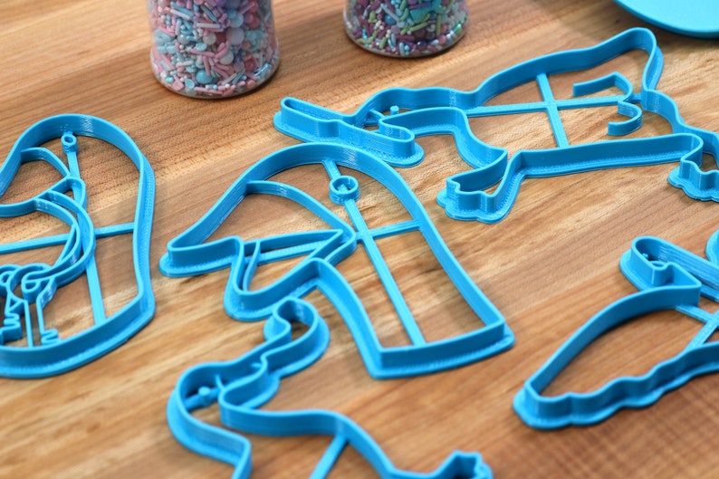 May include: A set of five blue plastic cookie cutters shaped like a flamingo, a mushroom, a bunny, a bird, and a butterfly. The cutters are on a wooden surface.