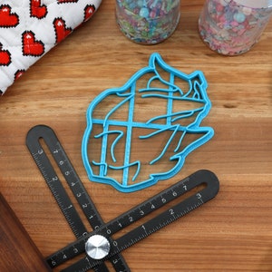 May include: A blue plastic cookie cutter shaped like a bear's head. The cutter is on a wooden surface with a ruler and a rolling pin.