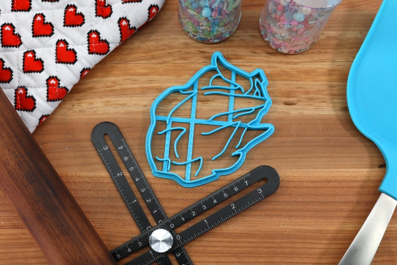 May include: A blue plastic cookie cutter shaped like a bear's head. The cutter is on a wooden surface with a ruler and a rolling pin.