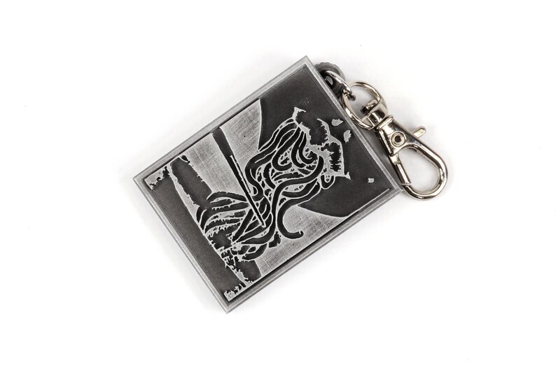May include: A silver metal keychain with a black and white image of a creature with tentacles. The keychain has a silver metal clasp.