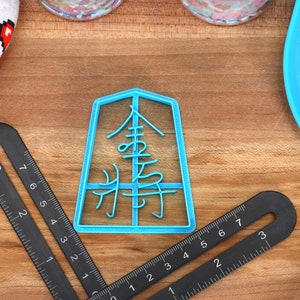 May include: A light blue plastic cookie cutter in the shape of Japanese characters, with a black ruler and a white and red heart-patterned cloth on a wooden surface. The cookie cutter features the Japanese characters for "Gold General".