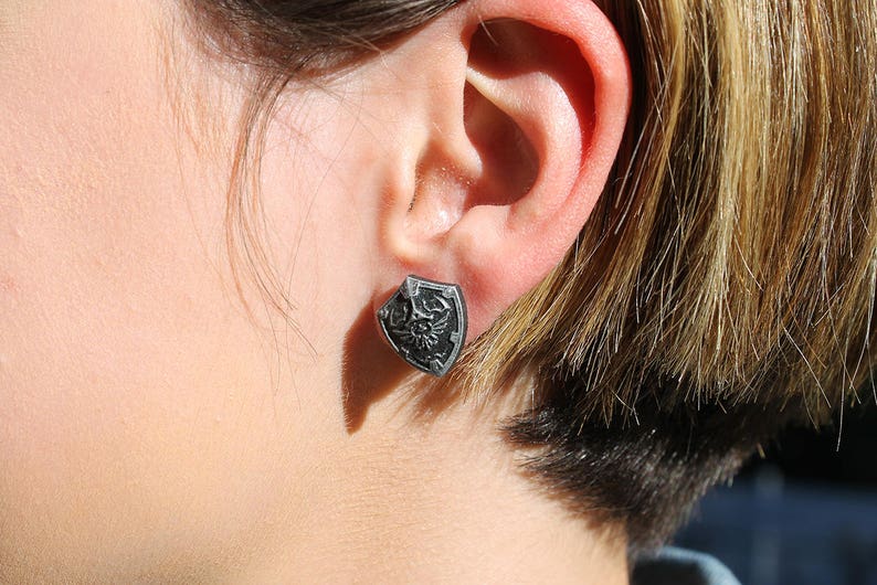 May include: A silver earring with a shield-shaped design. The shield has a black and white design with a stylized bird inside.