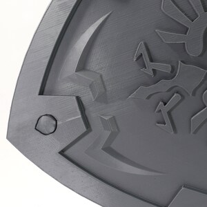 May include: A gray plastic shield with a raised design of a stylized bird with wings and a triangular symbol. The shield has a textured surface and a rounded edge.