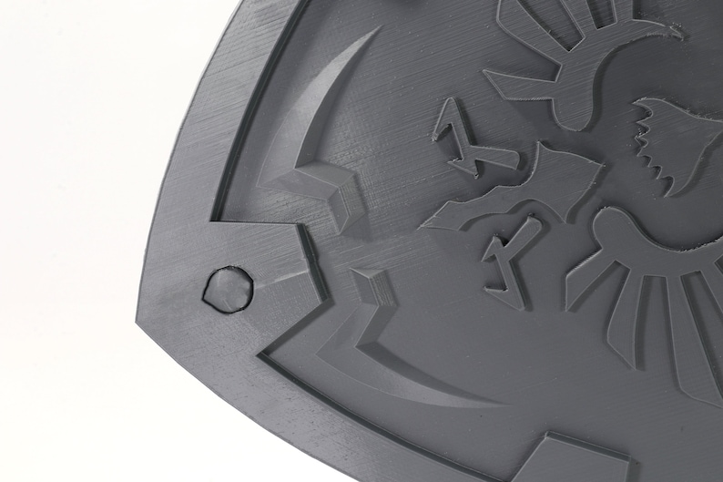 May include: A gray plastic shield with a raised design of a stylized bird with wings and a triangular symbol. The shield has a textured surface and a rounded edge.