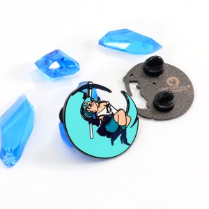 May include: A teal blue enamel pin featuring a cartoon character with blue hair sitting on a crescent moon, holding a scythe. The pin has a black metal backing with a butterfly design and the text "LOOT CAVE CO. - lootcaveco.com".