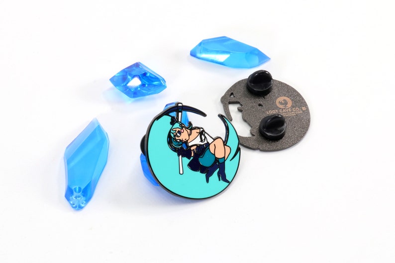 May include: A teal blue enamel pin featuring a cartoon character with blue hair sitting on a crescent moon, holding a scythe. The pin has a black metal backing with a butterfly design and the text "LOOT CAVE CO. - lootcaveco.com".