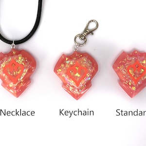 May include: Three pink resin heart-shaped charms with a glittery finish and a white heart design. The charms are shown on a black cord necklace, a silver keyring, and a stand.