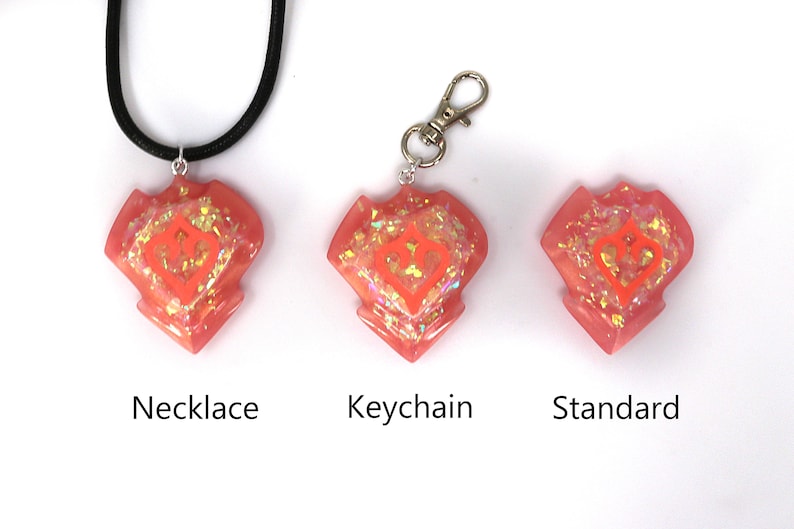 May include: Three pink resin heart-shaped charms with a glittery finish and a white heart design. The charms are shown on a black cord necklace, a silver keyring, and a stand.