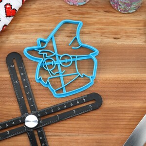 May include: A blue plastic cookie cutter shaped like a cartoon character wearing a hat and glasses. The cookie cutter is on a wooden surface.