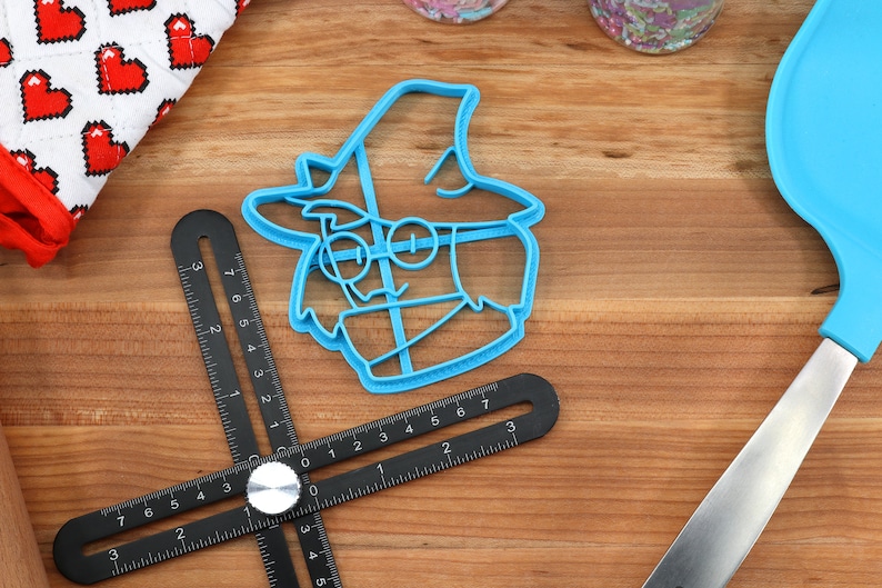 May include: A blue plastic cookie cutter shaped like a cartoon character wearing a hat and glasses. The cookie cutter is on a wooden surface.