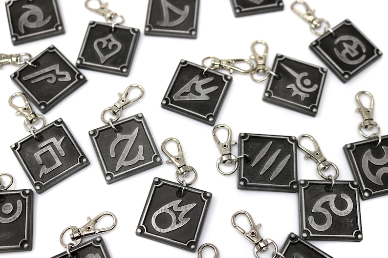May include: A set of 15 silver and black metal keychains with different symbols. Each keychain features a square-shaped charm with a unique symbol in a silver color on a black background.