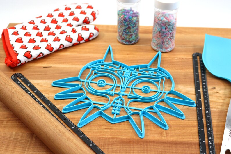 May include: A blue plastic cookie cutter shaped like the iconic Legend of Zelda Majora's Mask. The cutter is on a wooden surface with a rolling pin, a ruler, and two jars of sprinkles.