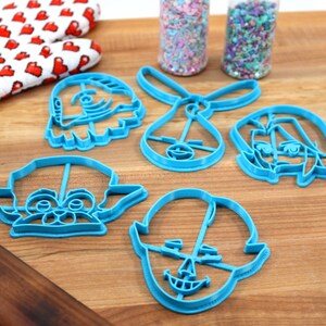 Pu&ograve; includere: Un set di cinque tagliabiscotti in plastica blu a forma di personaggi dei cartoni animati. I tagliabiscotti sono su una superficie di legno con un matterello e una spatola.