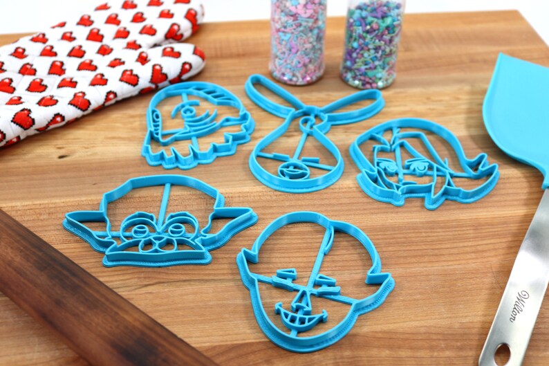 Pu&ograve; includere: Un set di cinque tagliabiscotti in plastica blu a forma di personaggi dei cartoni animati. I tagliabiscotti sono su una superficie di legno con un matterello e una spatola.