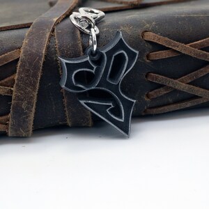 May include: A silver-colored metal keychain with a stylized design. The keychain is attached to a brown leather journal with a braided strap.