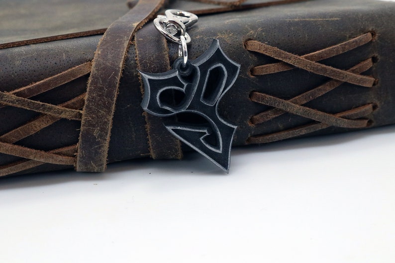 May include: A silver-colored metal keychain with a stylized design. The keychain is attached to a brown leather journal with a braided strap.