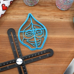 Puede incluir: Un cortador de galletas de pl&aacute;stico azul con forma de cara de dibujos animados con una amplia sonrisa y ojos grandes. El cortador est&aacute; sobre una superficie de madera con una regla y una esp&aacute;tula azul.