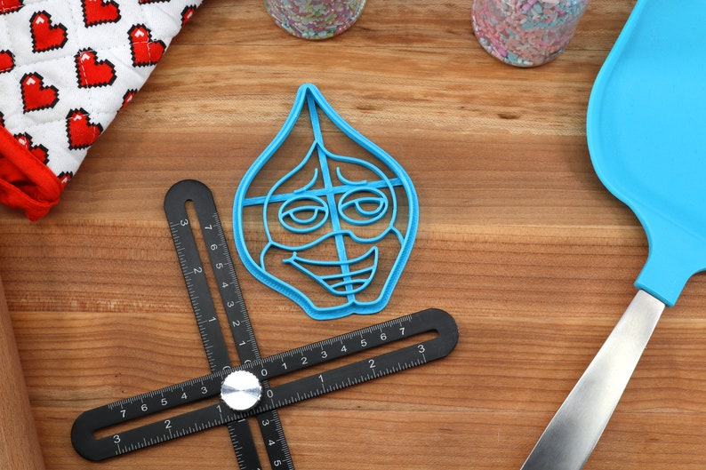 Puede incluir: Un cortador de galletas de pl&aacute;stico azul con forma de cara de dibujos animados con una amplia sonrisa y ojos grandes. El cortador est&aacute; sobre una superficie de madera con una regla y una esp&aacute;tula azul.