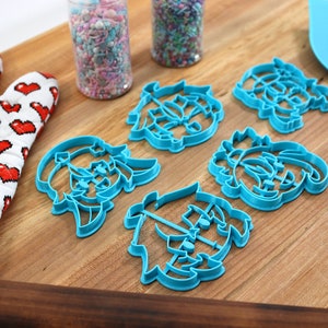 May include: Five blue plastic cookie cutters shaped like cartoon characters on a wooden cutting board. The cutters are arranged in a semi-circle with sprinkles in jars in the background.
