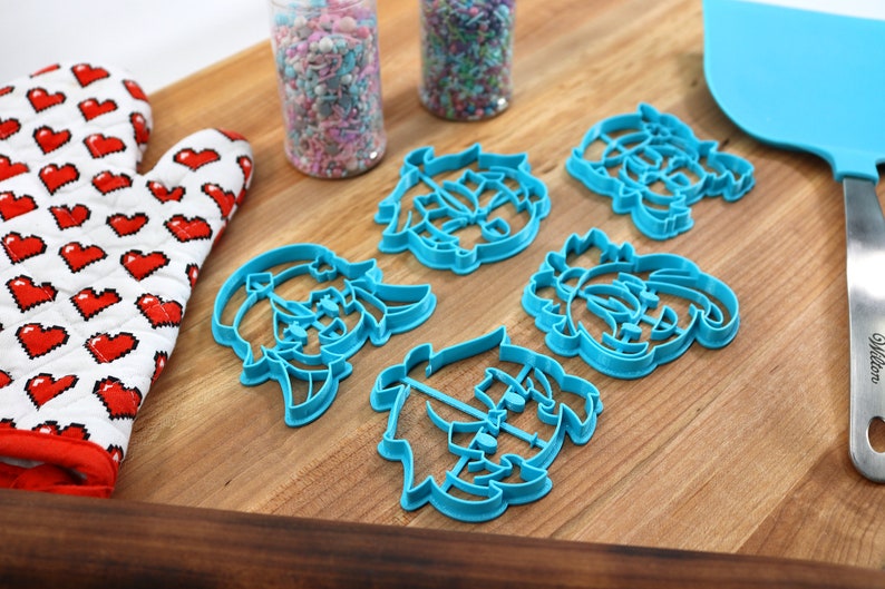 May include: Five blue plastic cookie cutters shaped like cartoon characters on a wooden cutting board. The cutters are arranged in a semi-circle with sprinkles in jars in the background.