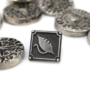 May include: A silver metal pin with a black square background and a silver leaf design. The pin is surrounded by several silver coins.